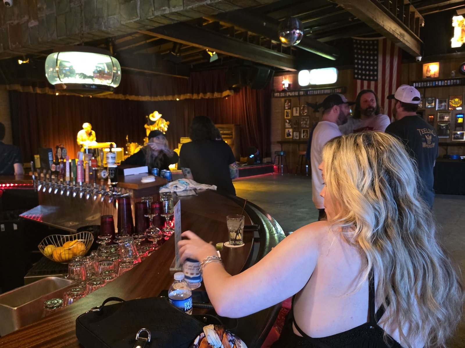 A woman sitting at a slow bar in a dimly lit live-music venue, with taps and barware in the foreground and a band performing on stage in the background.